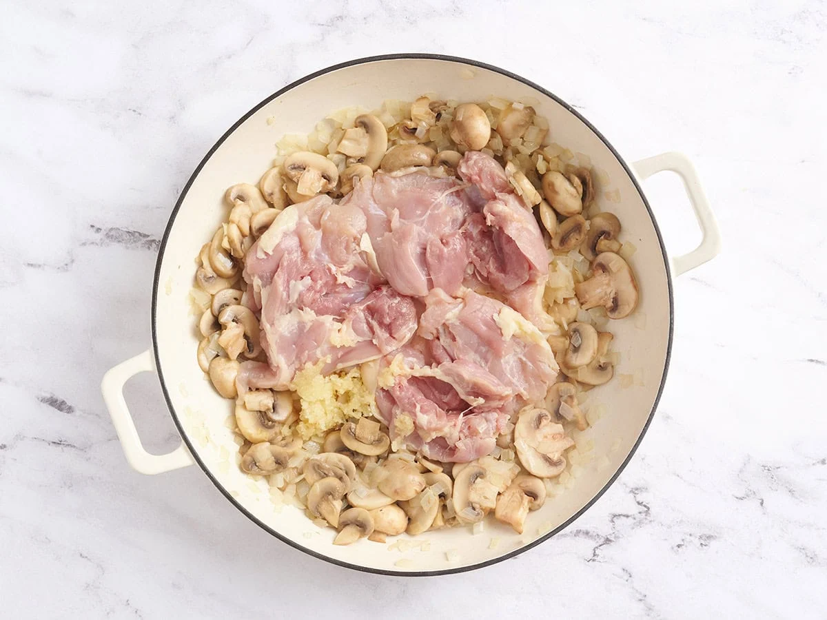 Cuisses de poulet ajoutées à une poêle avec des oignons et des champignons sautés.