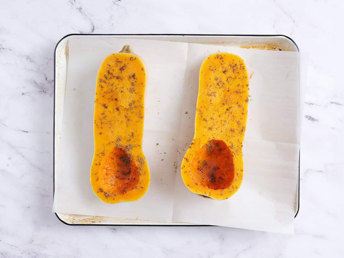 Moitiés de courge musquée assaisonnées sur une plaque à pâtisserie avant de les rôtir.