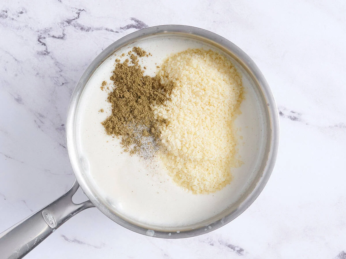 Assaisonnements et parmesan ajoutés à la sauce béchamel dans une casserole.