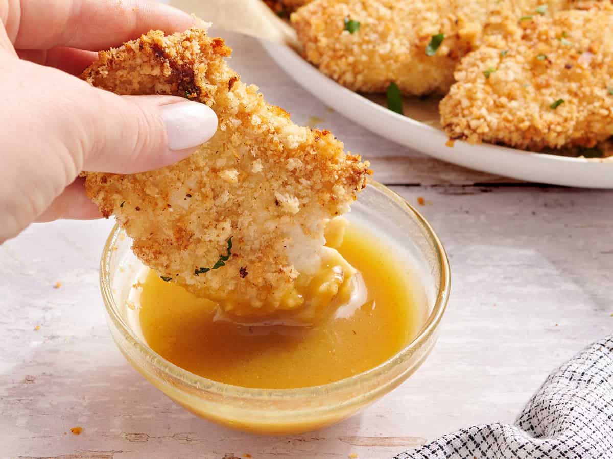 Une main trempant un demi-poulet tendre dans une sauce moutarde au miel.